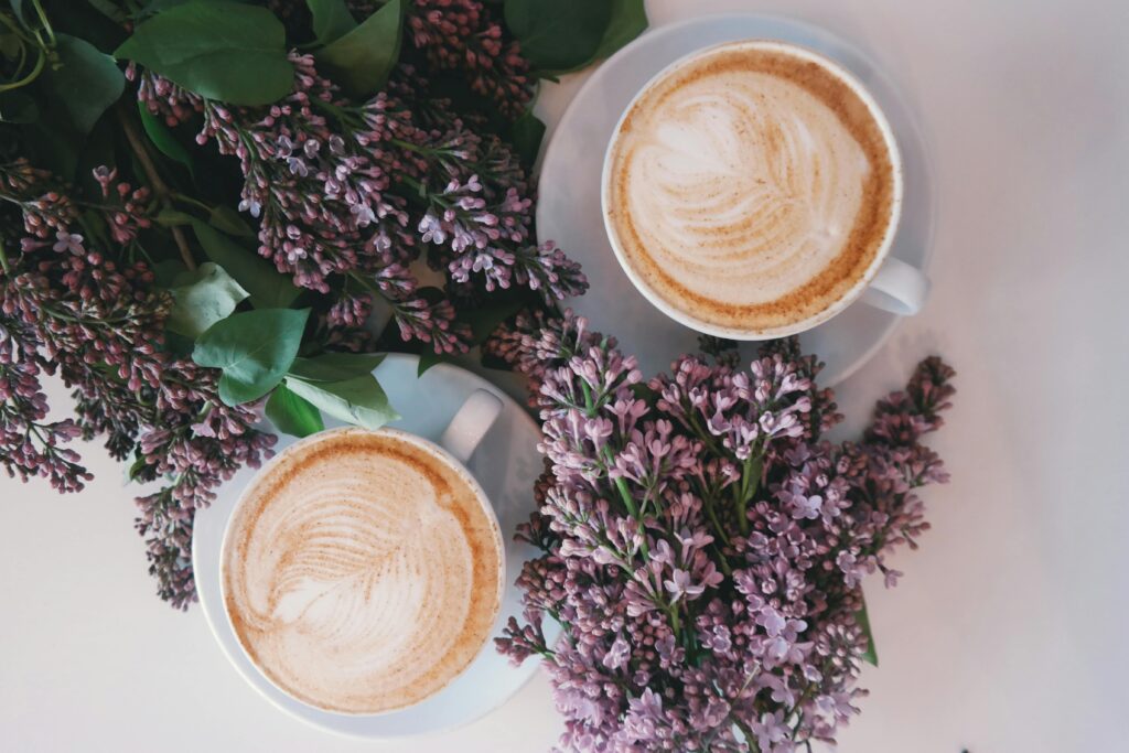 Two cups of latte art coffee are placed on a white surface next to bunches of purple lilac flowers, seen from an overhead view.