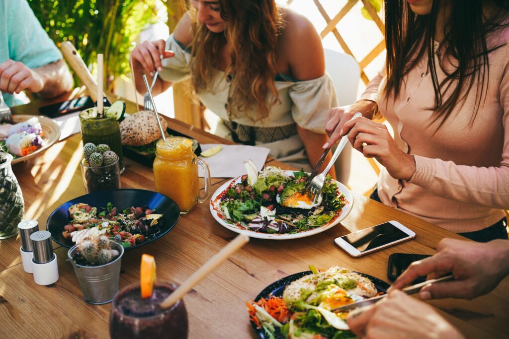 Close-up of a group of friends sharing a healthy brunch at a wooden outdoor table. The spread includes avocado toast with poached eggs, fresh salads, fruit smoothies in mason jars, and colorful spring rolls. Natural sunlight illuminates the vibrant food and casual dining setting.