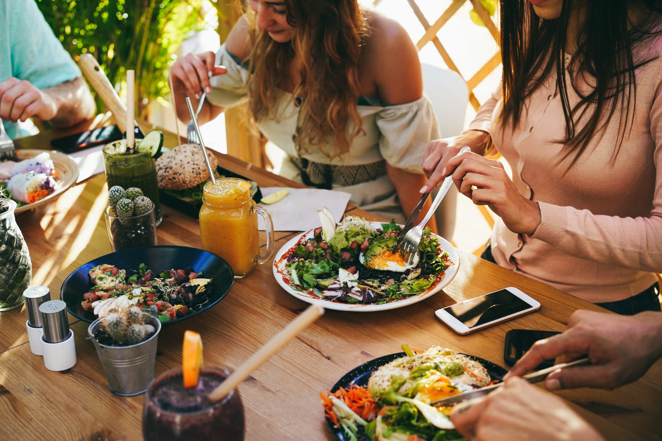 Close-up of a group of friends sharing a healthy brunch at a wooden outdoor table. The spread includes avocado toast with poached eggs, fresh salads, fruit smoothies in mason jars, and colorful spring rolls. Natural sunlight illuminates the vibrant food and casual dining setting.